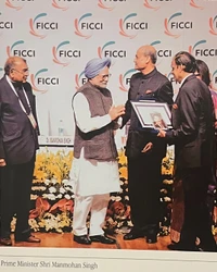 Ajay Rungta shakes hands with former Indian PM Manmohan Singh at a FICCI event. Another man presents a framed certificate.