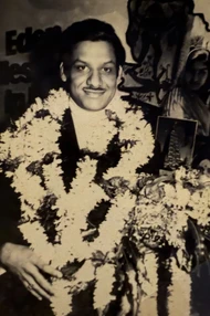 Black and white photo of Ajay Rungta (Dadu) smiling, wearing multiple flower garlands and holding a small award, looking distinguished.