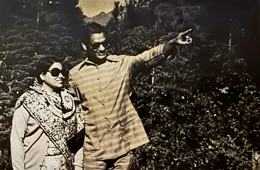 Vintage photo of Ajay Rungta (Dadu) and a woman, likely his wife 'Ma', both in sunglasses, outdoors. Dadu is pointing to the right.