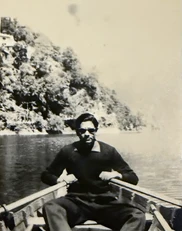 Vintage sepia photo of Ajay Rungta wearing sunglasses, rowing a boat on a calm lake with a forested shore in the background.