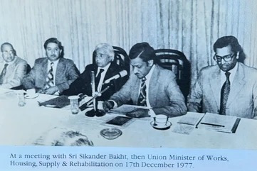 Black and white photo of Ajay Rungta (Dadu) at a formal meeting table with Sri Sikander Bakht and other officials in 1977.