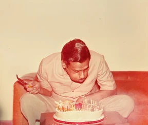 Ajay Rungta seated, leaning forward to blow out candles on a birthday cake during a celebration from his younger days.