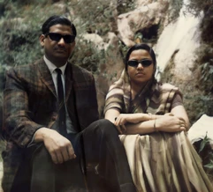 Ajay Rungta (Dadu) in a suit and his wife in a saree, both in sunglasses, seated outdoors. Vintage photo with natural rocky background.