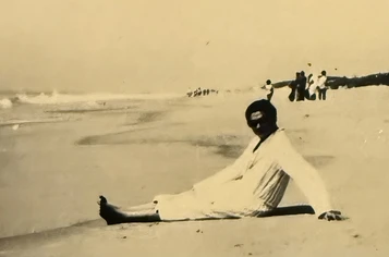 Vintage sepia photo of a young Ajay Rungta sitting relaxed on a sandy beach, with distant figures and the shoreline.