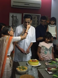 Sushila Buaji lovingly feeds cake to Ajay Rungta (Dadu) in a white kurta, with family including children around a table with a cake.