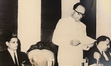 Vintage black and white photo: Ajay Rungta in a light suit, speaking at a podium, holding papers. Two men are seated listening.